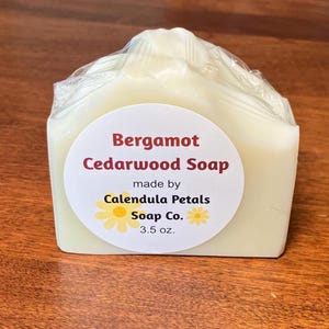 May include: A bar of Bergamot Cedarwood Soap, made by Calendula Petals Soap Co. The soap is wrapped in clear plastic and features a round label with the product name and company details. The bar weighs 3.5 oz.