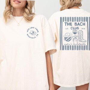 May include: Cream-colored t-shirt with a blue graphic on the front that says "With Love From Nashville TN." The back features a blue design with the text "THE BACH CLUB est. 2026" and illustrations of a cowboy hat, wine glass, and boots.