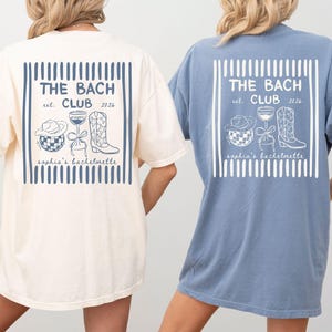 May include: Two oversized t-shirts, one cream and one blue, feature a graphic on the back with the text "THE BACH CLUB est. 2026" and illustrations of a cowboy hat, cocktail, and boot. The shirts are for a bachelorette party.