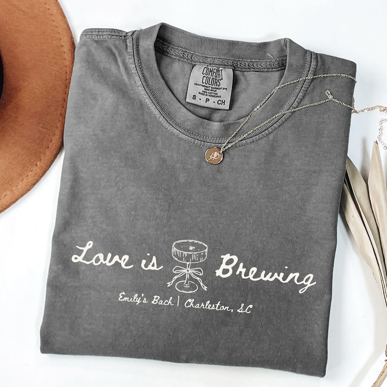 以下が含まれることがあります： 「Love is Brewing」の文字とカクテルのグラフィックがプリントされたグレーのComfort Colors Tシャツ。 「Emily's Bach | Charleston, SC」の文字もプリントされています。 金色のネックレスと茶色の帽子も画像にあります。