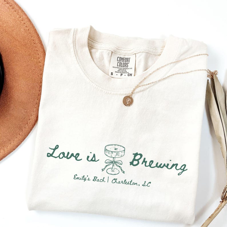 以下が含まれることがあります： 「Love is Brewing」の緑色の文字とカクテルテーブルのグラフィックがプリントされた、クリーム色のTシャツ。「Emily's Bach | Charleston, SC」の文字が下にあります。金のネックレスと茶色の帽子も写っています。