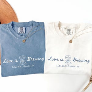 以下が含まれることがあります： 「Love is Brewing」の文字と、樽とテーブルのグラフィックが描かれた、青とオフホワイトの2枚のTシャツ。テキスト「Emily's Bach | Charleston, SC」が下にあります。各シャツには金のネックレスが付いています。