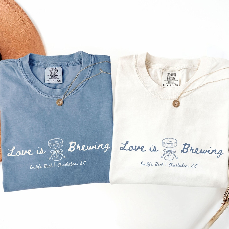 以下が含まれることがあります： 「Love is Brewing」の文字と、樽とテーブルのグラフィックが描かれた、青とオフホワイトの2枚のTシャツ。テキスト「Emily's Bach | Charleston, SC」が下にあります。各シャツには金のネックレスが付いています。