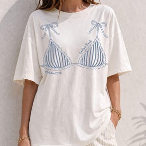 May include: Off-white t-shirt featuring a blue and white striped bikini top design. The design includes bows, scalloped edges, and the text "Emily's Bach" and "Malibu, 2026". The shirt has short sleeves and a relaxed fit.
