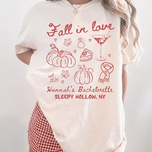 May include: Cream-colored t-shirt with a red graphic design. The design includes the text "Fall in love", pumpkins, maple leaves, a ring, a cocktail, a slice of pie, and a scarf. The text "Hannah's Bachelorette SLEEPY HOLLOW, NY" is also included.