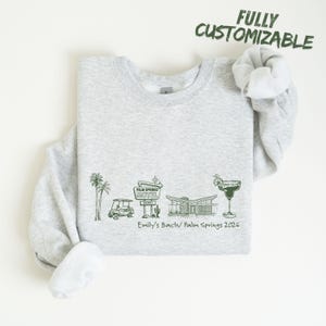May include: A light gray sweatshirt with a green graphic design featuring palm trees, a golf cart, a motel sign, a mid-century modern house, and a margarita glass. The text "Emily's Bach/ Palm Springs 2026" is below the graphic. The words "FULLY CUSTOMIZABLE" are above the graphic.