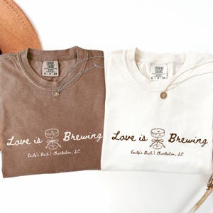 Può includere: Due magliette, una marrone e una bianca, con la frase "Love is Brewing" e una grafica di un tavolo con un fiocco. Ogni maglietta ha una collana d'argento con un ciondolo. Il testo "Emily's Bach | Charleston, SC" è sotto.