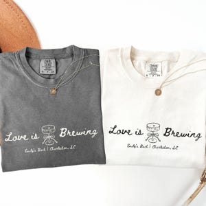 以下が含まれることがあります： 「Love is Brewing」の文字と醸造装置のグラフィックがプリントされた、チャコールグレーとオフホワイトの2枚のComfort Colors Tシャツ。各シャツには金のペンダントネックレスが付いています。「Emily's Bach | Charleston, SC」の文字が下にあります。