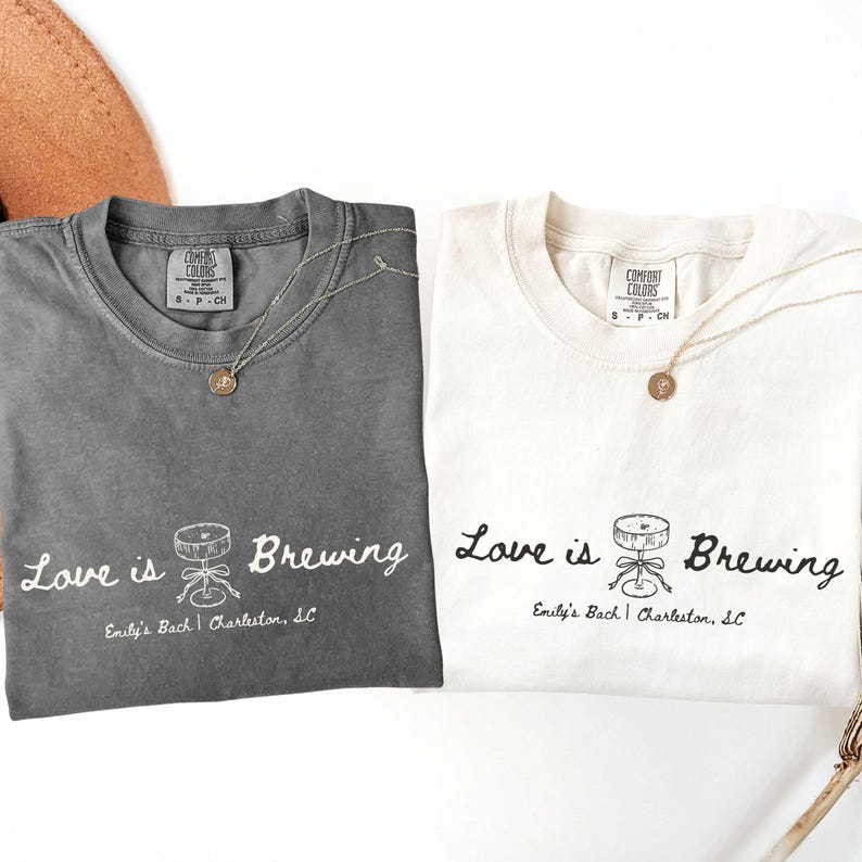 以下が含まれることがあります： 「Love is Brewing」の文字と醸造装置のグラフィックがプリントされた、チャコールグレーとオフホワイトの2枚のComfort Colors Tシャツ。各シャツには金のペンダントネックレスが付いています。「Emily's Bach | Charleston, SC」の文字が下にあります。