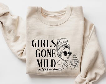 May include: A beige sweatshirt with the text "Girls Gone Mild" and a drawing of a woman with a towel on her head, sunglasses, and a cup in her hand. The text "est. 2025" and "emily's bachelorette" are also included.