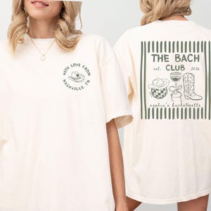 May include: Cream-colored t-shirt with a front graphic that says "With Love From Nashville TN" and a back graphic that says "The Bach Club est. 2026" with illustrations of a cowboy hat, wine glass, and boots. Sophia's bachelorette.
