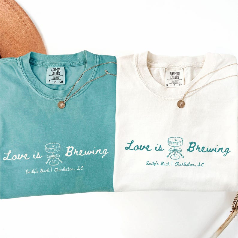 以下が含まれることがあります： 「Love is Brewing」の文字とカクテルシェーカーのグラフィックがプリントされた、ティールとオフホワイトの2枚のTシャツ。各シャツにはペンダント付きのゴールドネックレスが付いています。「Emily's Bach | Charleston, SC」の文字が下にあります。