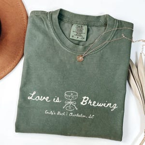 以下が含まれることがあります： 「Love is Brewing」の文字と樽のイラストがプリントされたオリーブグリーンのTシャツ。下には「Emily's Bach | Charleston, SC」の文字。金色のペンダント付きネックレスがTシャツの上に置かれています。