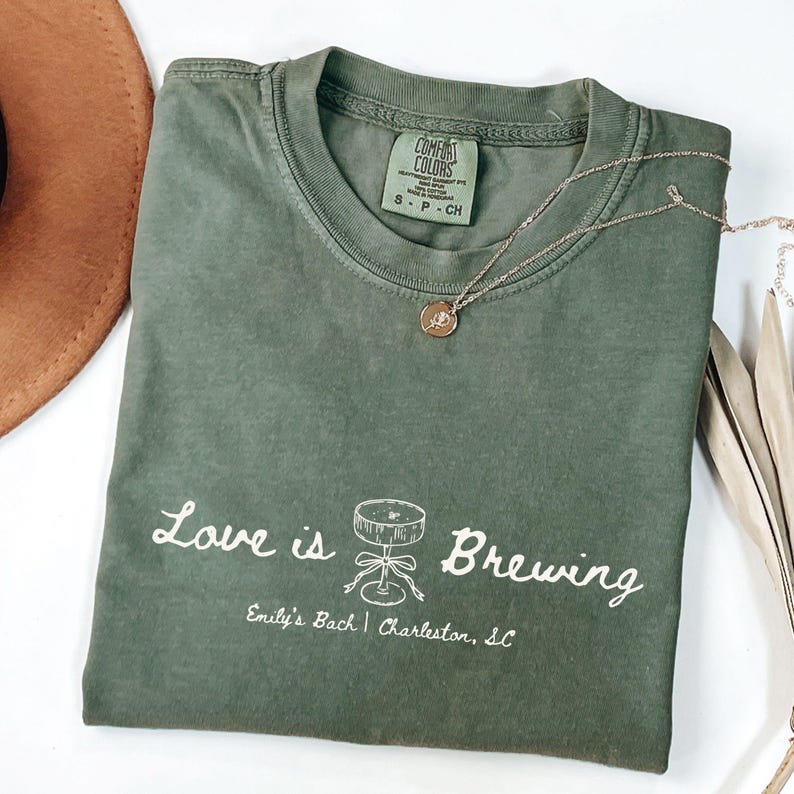 以下が含まれることがあります： 「Love is Brewing」の文字と樽のイラストがプリントされたオリーブグリーンのTシャツ。下には「Emily's Bach | Charleston, SC」の文字。金色のペンダント付きネックレスがTシャツの上に置かれています。