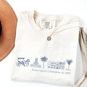 May include: Off-white t-shirt featuring a blue illustration of a carriage, pineapple fountain, palm tree, buildings, and a cocktail. The text "Emily's Bach/ Charleston, SC 2025" is printed below. A gold necklace is draped on the shirt.