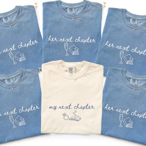 May include: Five blue t-shirts and one cream t-shirt. The blue shirts feature white text reading 'her next chapter' with a champagne flute and book graphic. The cream shirt features dark text reading 'my next chapter' with a ring and book graphic.