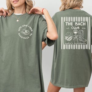 May include: Olive green t-shirt with a white graphic on the front that says "With Love From Nashville TN". The back of the shirt has a white graphic that says "The Bach Club est. 2026" with illustrations of a cowboy hat, disco ball, wine glass, and cowboy boot.