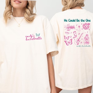May include: Two white t-shirts with pink and teal text and graphics. The front of one shirt says "emily's bachelorette" and the back of the other shirt says "He Could Be the One" with a graphic of a disco ball, sunglasses, boots, a guitar, butterflies, and a woman with a microphone.