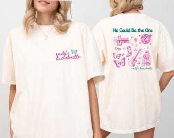 May include: Two white t-shirts with pink and teal text and graphics. The front of one shirt says "emily's bachelorette" and the back of the other shirt says "He Could Be the One" with a graphic of a disco ball, sunglasses, boots, a guitar, butterflies, and a woman with a microphone.
