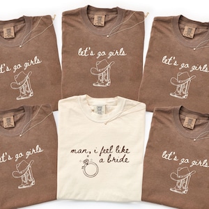 May include: Several brown and cream-coloured t-shirts are displayed. Most feature the text "let's go girls" with a cowboy hat and boot design. One shirt says "man, I feel like a bride" with a ring graphic. Each shirt has a necklace.
