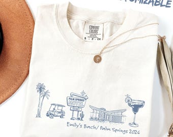 Può includere: T-shirt color crema con grafica blu di un motel di Palm Springs, palme e un cocktail. Il testo "Emily's Bach/ Palm Springs 2026" è stampato sotto. Una collana d'oro è drappeggiata sulla maglietta.
