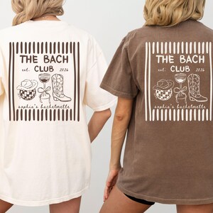May include: Two oversized t-shirts, one cream and one brown, feature a graphic on the back. The graphic includes the text "THE BACH CLUB est. 2026" and illustrations of a cowboy hat, wine glass, and cowboy boot. The text "sophia's bachelorette" is also included.