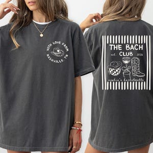 May include: Dark gray t-shirt with white graphic designs. The front features a circular design with a cowboy hat and the text "With Love From Nashville, TN." The back displays "THE BACH CLUB est. 2026" with related illustrations.