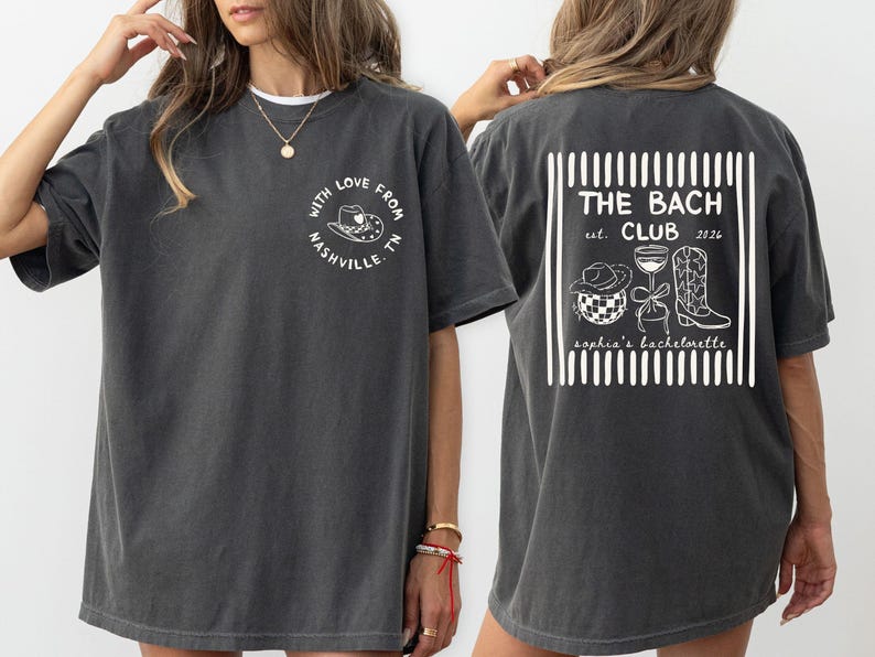 May include: Dark gray t-shirt with white graphic designs. The front features a circular design with a cowboy hat and the text "With Love From Nashville, TN." The back displays "THE BACH CLUB est. 2026" with related illustrations.