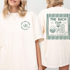 May include: Cream-colored t-shirt with green text and graphics. The front features a circular design with a cowboy hat and the text "With Love From Nashville TN." The back displays "THE BACH CLUB est. 2026" with related illustrations.