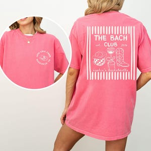 May include: A pink t-shirt with white text and graphics. The back of the shirt features a design with the words "THE BACH CLUB" and the establishment year "2026". The front of the shirt has the words "WITH LOVE FROM NASHVILLE TN".