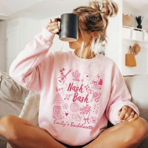 May include: A light pink sweatshirt with the words "Nash Bash" and "Emily's Bachelorette" in a pink script. The sweatshirt features pink illustrations of a guitar, cactus, cake, boots, and other celebratory icons. A black mug is held up.