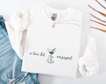May include: White sweatshirt with the text "a tini bit engaged" and a green martini glass graphic. A silver necklace with a small pendant rests on the sweatshirt. The sweatshirt is folded with a pair of blue jeans.