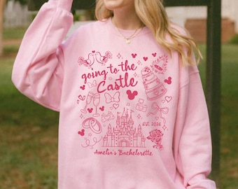 May include: A light pink long-sleeve sweatshirt featuring a dark pink graphic print with the text "going to the Castle", "Est. 2026", and "Amelia's Bachelorette". The design includes illustrations of a castle, wedding cake, rings, champagne flutes, and bows.