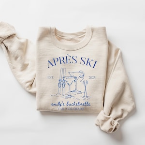 May include: A beige sweatshirt with a blue graphic design that says "Aprés Ski" with a ski design and the text "emily's bachelorette Vail, Colorado"
