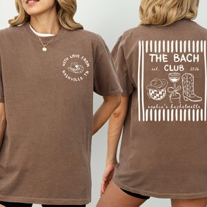 May include: Brown t-shirt with white text and graphics. Front reads "WITH LOVE FROM NASHVILLE, TN" in a circle. Back features "THE BACH CLUB est. 2026" with a cowboy hat, disco ball, wine glass, and boot.