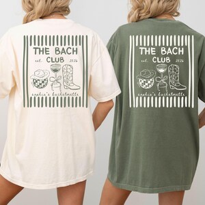 May include: Two short-sleeved t-shirts, one ivory and one moss green, feature a graphic design on the back. The design includes the text "THE BACH CLUB est. 2026" within a rectangular frame, along with illustrations of a cowboy hat, a disco ball, a wine glass, and a cowboy boot. The text "sophia's bachelorette" is also included.