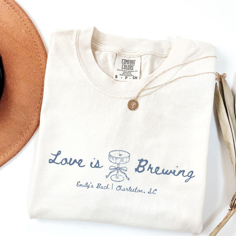 以下が含まれることがあります： クリーム色のTシャツ。青い文字で「Love is Brewing」とカクテルのイラストが描かれています。「Emily's Bach | Charleston, SC」の文字も。金色のネックレスが折りたたまれたTシャツの上に置かれています。
