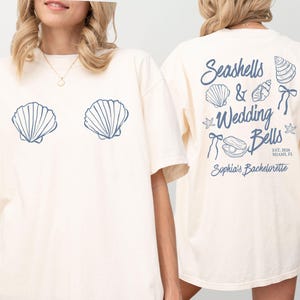May include: Cream-colored t-shirt with a seashell design on the front and back. The front features two blue seashell graphics. The back displays the text "Seashells & Wedding Bells" with seashell and bow illustrations, and "Sophia's Bachelorette" below.