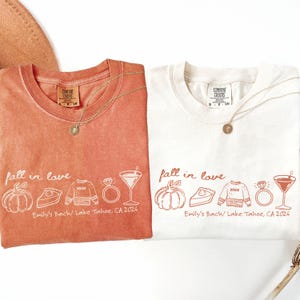 May include: Two crewneck t-shirts, one terracotta and one white, featuring a graphic print with the text "fall in love" and "Emily's Bach/ Lake Tahoe, CA 2026". The design includes line art of a pumpkin, pie slice, sweater, ring, and martini glass.
