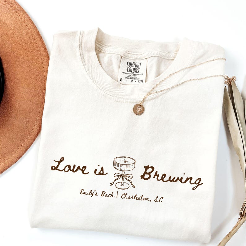 以下が含まれることがあります： 「Love is Brewing」の文字とカクテルのイラストが描かれたクリーム色のTシャツ。金のネックレスと茶色の帽子も見える。テキスト「Emily's Bach | Charleston, SC」がグラフィックの下にあります。