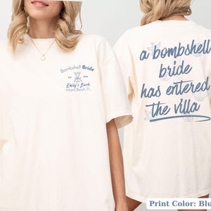 May include: An off-white short-sleeve t-shirt with blue text. The front features a martini glass icon and the text Bombshell Bride, Est 2024, Emily's Bach, Miami Beach, FL. The back displays the text a bombshell bride has entered the villa.