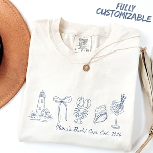May include: A cream-colored t-shirt featuring blue line art of a lighthouse, a bow, a lobster, a seashell, and a cocktail glass. Below the graphics, the text reads Olivia's Bach/ Cape Cod, 2026. The shirt has a Comfort Colors brand label.