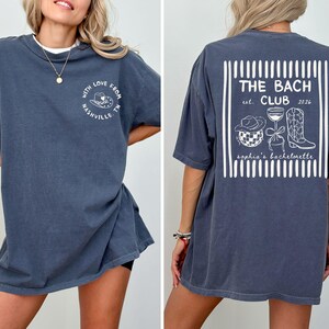 May include: Dark blue t-shirt with white text and graphics. The front says "With Love From Nashville, TN" around a cowboy hat. The back says "THE BACH CLUB est. 2026" with a hat, wine glass, and boots.