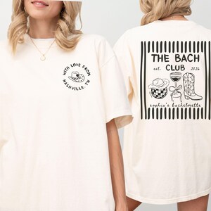 May include: Cream-colored t-shirt with black graphic designs. The front features a cowboy hat and the text "WITH LOVE FROM NASHVILLE, TN." The back displays "THE BACH CLUB est. 2026" with a hat, wine glass, and boots.