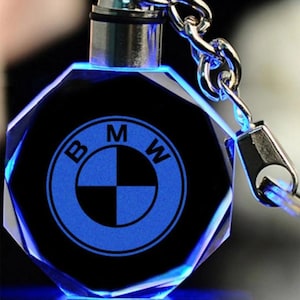 Könnte beinhalten: Ein schwarzer und blauer Kristall-Schlüsselanhänger mit dem BMW-Logo. Der Schlüsselanhänger hat ein facettiertes Design und eine silberne Metallkette und einen Verschluss. Das BMW-Logo ist blau beleuchtet, und die Kanten sind mit blauem Licht versehen.