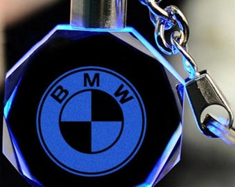 May include: A black and blue crystal keychain featuring the BMW logo. The keychain has a faceted design and a silver metal chain and clasp. The BMW logo is illuminated in blue, and the edges are highlighted with blue light.
