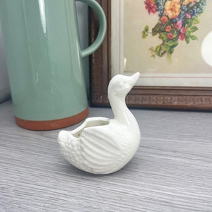 Puede incluir: Macetero de cerámica blanca en forma de cisne con superficie texturizada. El cisne tiene un cuello largo y una pequeña abertura en la parte posterior para plantar.