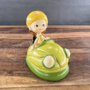 May include: A ceramic figurine of a girl wearing a yellow helmet and a yellow dress driving a green bumper car.