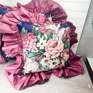 May include: A cushion with a navy blue floral design, showcasing pink, white, and red flowers. It's framed by a ruffled, deep pink fabric. This cushion is part of a bedding set.