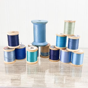May include: A collection of vintage wooden spools of thread in various shades of blue, teal, and green. The spools are arranged on a reflective surface, showcasing the different colors and textures of the thread.